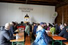 Viele Menschen sitzen in einer Maschinenhalle an Biertischen, ein Mann steht vorne am Rednerpult
