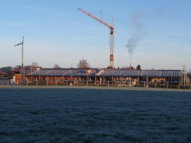 Das Foto zeigt die aktuelle Baustelle für den neuen Kälberstall in Achselschwang von Süden. Zu sehen ist das Stallgebäude in seiner vollen Länge mit einem davor errichteten Gerüst sowie die teilweise schon vollflächig montierten PV-Module.